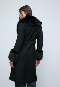 Donna che indossa un cappotto nero con cintura e bordo in pelliccia su collo e polsini, mostrata di spalle su uno sfondo neutro.