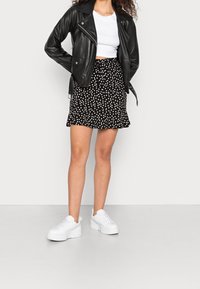 Schwarze Leder-Bikerjacke über einem weißen Crop-Top, kombiniert mit einem schwarz-weißen Pünktchen-Minirock und klobigen weißen Sneakers.