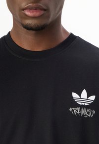 Melnas kokvilnas t-krekls ar īsām piedurknēm, ar baltu Adidas logotipu ar trim lapām un grafikām teksta tuvu logotipam, gluda tekstūra un apaļš kakla izgriezums.