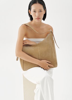 Žena s krátkymi čiernymi vlasmi, oblečená v bielom odeve, nesie veľkú hnedú semišovú kabelku cez plece na bielom pozadí.