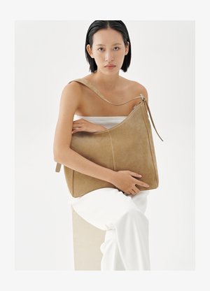 Sieviete ar īsiem melniem matiem baltā apģērbā, turot lielu gaišbrūnu zamšādas plecu somu pret baltu fonu.