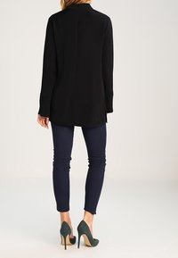 Blusa negra de manga larga con cremallera en la parte posterior, con aberturas laterales; combinada con pantalones estrechos azules marino y tacones verdes. Textura de tela suave.
