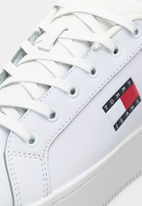 Biele tenisky s bielymi šnúrkami a sieťovinovým jazykom, s logom Tommy Jeans na boku nad podrážkou.