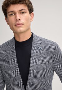 Blazer con un motivo houndstooth geometrico in grigio e blu navy, dotato di una vestibilità su misura, rever a punta e una spilla blu sul lato sinistro.