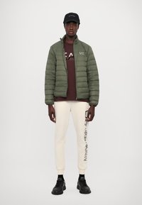 Puffer jacket in olive green, zip-up design. Underneath, a maroon shirt with "EA7" logo. Cream sweatpants with side branding. Black sneakers.