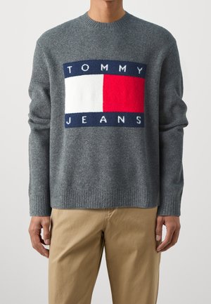 Mężczyzna w szarym swetrze z dużym logo Tommy Jeans w kolorach niebieskim, białym i czerwonym z przodu, w połączeniu z beżowymi spodniami, na jednolitym tle.
