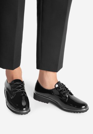 Błyszczące czarne buty na niskim obcasie z sznurowadłami, noszone z czarnymi, krótkimi spodniami, odsłaniającymi kostki, na białym tle.