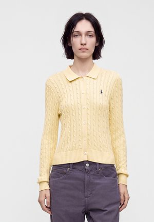 Femme aux cheveux courts et foncés, portant un cardigan jaune en maille torsadée boutonné et un pantalon gris, debout devant un fond uni.
