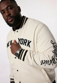 Krämfärgad varsityjacka med svart randig ribbad krage och muddar. Har "NEW YORK" broderat fram och skriptdetalj längs ärmen.