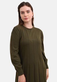 Olivgrünes Strickkleid mit langen Ärmeln, gerippter Textur, rundem Ausschnitt und vertikalen Nähten, die eine figurbetonte Silhouette schaffen.