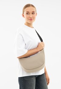 Beige műbőr válltáska ívelt formával, fekete állítható pánttal és sima felülettel. Az elején dombornyomott logóval rendelkezik.