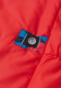 Rode pufferjas met een blauw en zwart patroon stofpatch. Heeft een zwarte drukknoop en een gladde, glanzende textuur.