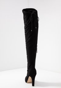 Bottes noires à talons hautes, au-dessus du genou, avec une texture lisse, présentant un laçage sur le côté, une fermeture éclair à l'arrière et un design à bout pointu.