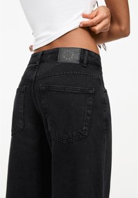 Gros plan sur une personne soulevant un haut blanc, portant un jean en denim noir taille haute avec des poches arrière et une étiquette sur la ceinture.