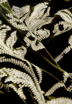 Samtstoff in tiefgrün mit aufwendiger beiger Stickerei, die florale Muster und strukturierte Akzente aufweist. Weiche und luxuriöse Oberfläche.