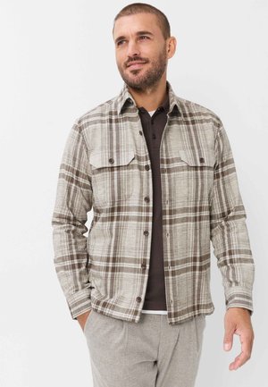 Mand med kort hår og skæg iført en beige og brun ternet skjorte med knapper over en mørk poloshirt og lysegrå bukser, stående mod en hvid baggrund.