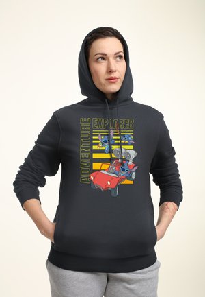 Kobieta w czarnej bluzie z kapturem z napisem „Adventure Explorer” i postaciami z kreskówek jadącymi czerwonym samochodem, pozująca z rękami w kieszeniach.