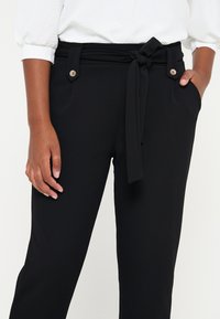 Černé kalhoty na míru s hladkou texturou, detaily vázacího pásku, dvě boční kapsy a dekorativní knoflíkové akcenty. Nosič má na sobě bílý top.