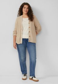 Beige gebreide cardigan met knopen, gedragen over een licht gestreept shirt. Gekozen in combinatie met blauwe jeans en multi-kleurige sneakers.