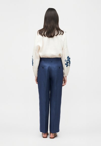 Γυναίκα όρθια με την πλάτη στην κάμερα, φοράει κρεμ μπλούζα με μπλε λουλουδάτη κεντητική και ψηλόμεσο μπλε παντελόνι με κουμπωτές πίσω τσέπες.