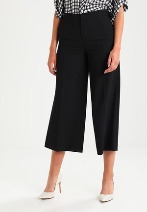 Pantalons larges noirs en tissu lisse, avec une taille haute et des plis, associés à des escarpins blancs à bouts pointus. Chemise à carreaux en arrière-plan.