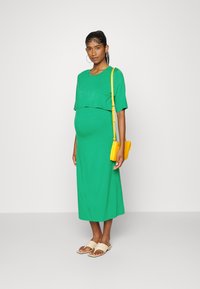 Vestuário verde de manga curta com uma silhueta fluida, acompanhado de uma pequena bolsa amarela e sandálias de slide bege.