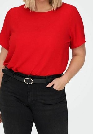 Frau trägt ein leuchtend rotes kurzärmeliges Oberteil, in hoch taillierte schwarze Jeans gesteckt, mit schwarzem Gürtel und goldener Schnalle, Hand in der Tasche.