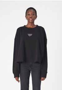 Black cropped sweatshirt with long sleeves, featuring a white embroidered "tommy jeans" logo on the chest. Paired with dark jeans.