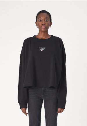 Felpa nera a maniche lunghe e taglio corto, con un logo ricamato bianco "tommy jeans" sul petto. Abbinata a jeans scuri.