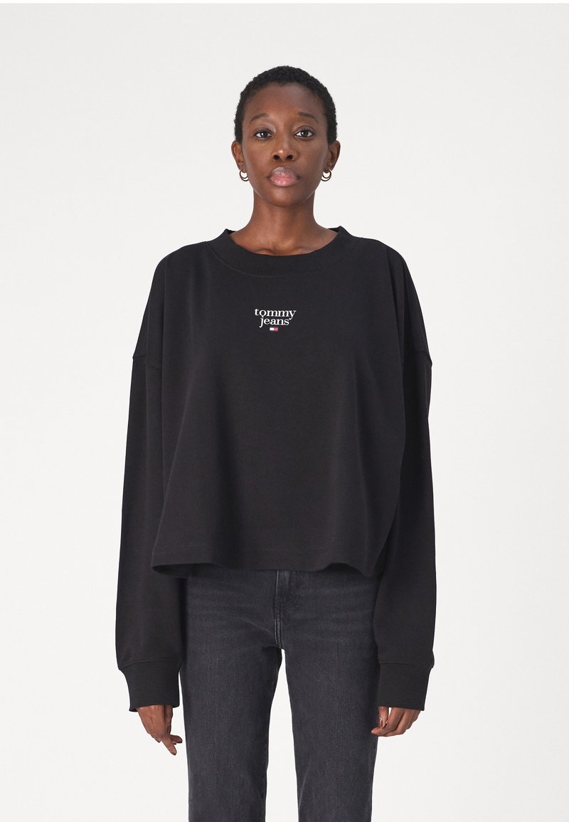 Black cropped sweatshirt with long sleeves, featuring a white embroidered "tommy jeans" logo on the chest. Paired with dark jeans.