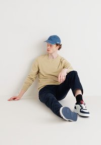 Sweatshirt beige, pantalon marine et casquette bleue. Les chaussures sont marines avec des accents blancs. La texture est lisse avec une coupe décontractée et une pose relaxée.