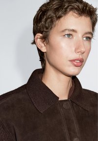Person med kort krøllet hår iført en mørkebrun jakke med en ribbet krave, der ser let mod højre foran en lys ensfarvet baggrund.