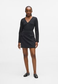 Abito nero con maniche lunghe, scollo a V, vita aderente e due tasche frontali. Tessuto liscio con una leggera lucentezza, abbinato a mocassini neri.