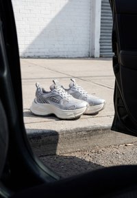 Baskets blanches et grises placées sur un trottoir ensoleillé à côté d'une porte de voiture ouverte avec un mur en briques blanches en arrière-plan.