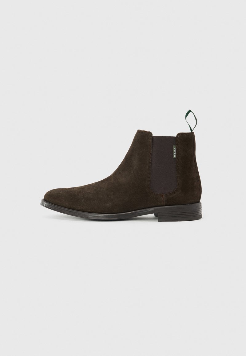 Bottine Chelsea en daim marron foncé avec panneau latéral élastique noir, talon bas empilé et languette de préhension verte à l'arrière sur fond clair.