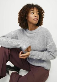 Pull tricot gris clair avec des coutures blanches décoratives le long des manches, associé à un pantalon bordeaux et des chaussures noires. Texture douce visible.