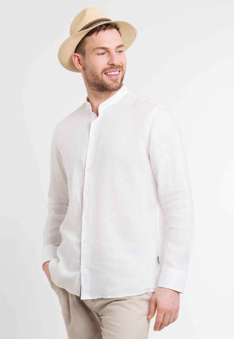 Chemise en lin blanche avec un col chinois, des manches longues et une fermeture à boutons. Portée avec un pantalon beige et un chapeau en paille léger.
