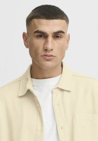 Beige T-shirt met textuur en kraag, met een borstzak, gelaagd over een witte ronde hals T-shirt.