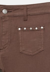 Jeans din denim maro, cu un buzunar posterior prevăzut cu cinci tije de argint, închidere cu nasture metalic și cusături contrastante.
