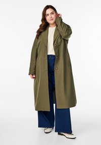 Olijfgroene trenchcoat met een gestructureerde silhouet, enkelvoudige knoopsluiting en geborduurde accenten op de mouwen, gecombineerd met wijde spijkerbroeken.
