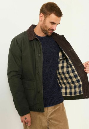Ein Mann trägt eine grüne Jacke mit braunem Kragen und zeigt das blau-beige karierte Futter innen in der Jacke.
