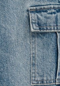 Gai zila džinsa auduma ar teksturētu aušanu, kuram ir krūšu kabata un kontrastējoša šuve. Virsma izskatās nolietota.