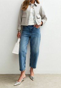Veste grise courte avec poches, haut blanc, jeans taille haute bleus, chaussures à talons argentées, tenant un sac à main blanc. Textures lisses et lignes épurées.