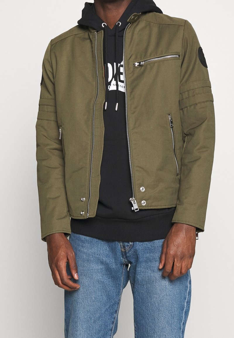 Man som bär en olivgrön zip-up jacka över en svart huva med vit tryck och blå jeans, visad från halsen till mitten av låren, stående mot en neutral bakgrund.