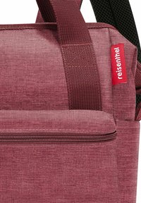Bolsa de tela roja con un acabado texturizado, que presenta un cierre superior, asas resistentes y una etiqueta lateral con el nombre de la marca.