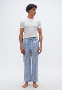 Νεαρός άνδρας που στέκεται ξυπόλητος, φορώντας λευκό μπλουζάκι και παντελόνι πιτζάμας σε απαλό μπλε καρό, κοιτάζοντας προς τα δεξιά του.