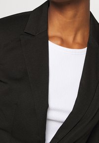 Blazer noir côtelé avec un col cranté, associé à un débardeur blanc côtelé. Accent sur la texture, le matériau et le design superposé.