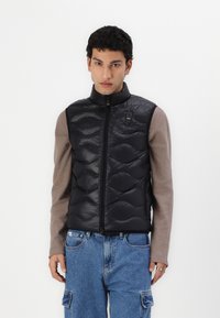 Svart puffer vest med quiltet tekstur, glidelås og høy krage. Brukt over en brun langermet skjorte, sammen med blå jeans.