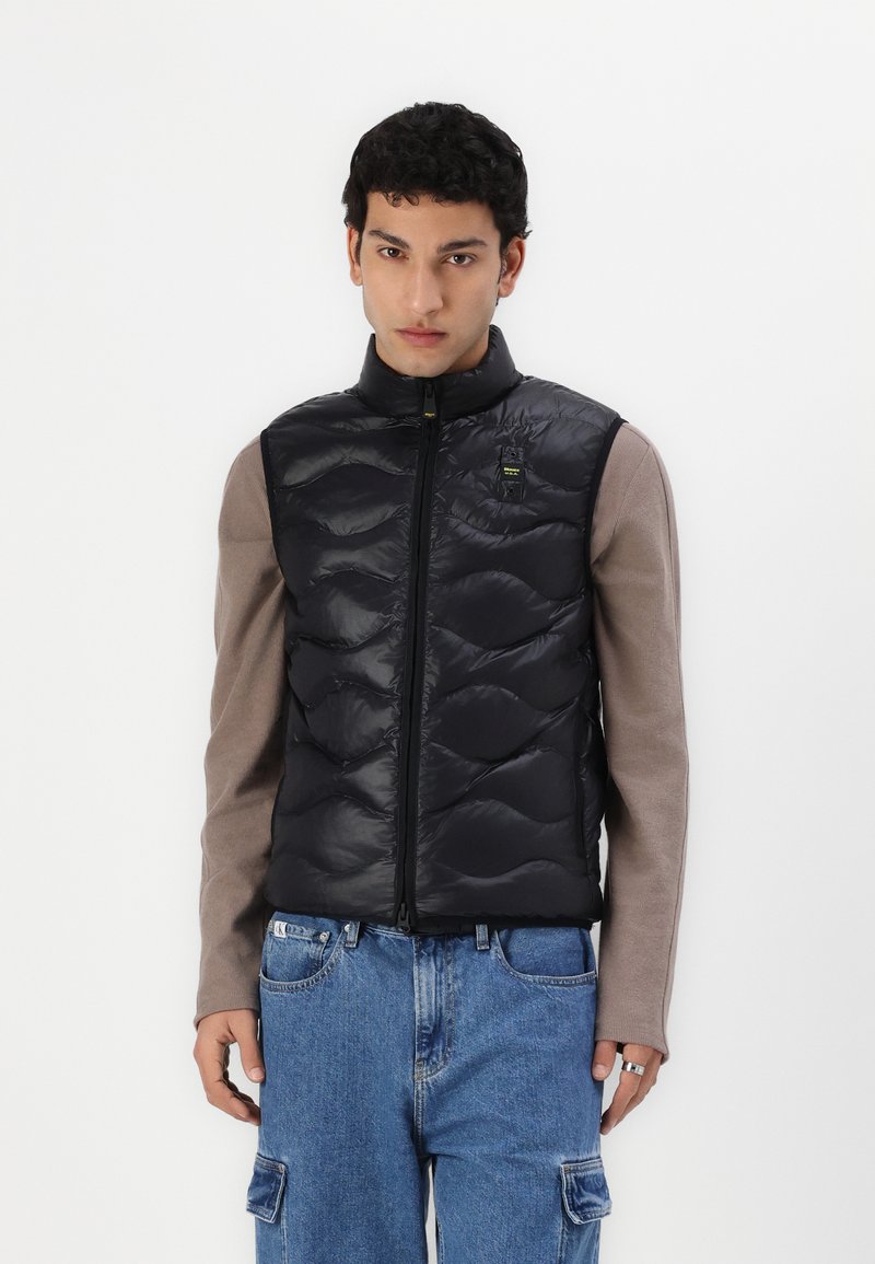 Svart puffer vest med quiltet tekstur, glidelås og høy krage. Brukt over en brun langermet skjorte, sammen med blå jeans.