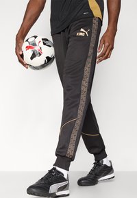 Calças desportivas pretas com detalhes dourados e risca lateral geométrica, combinadas com chuteiras pretas. Uma bola de futebol é segurada numa das mãos.